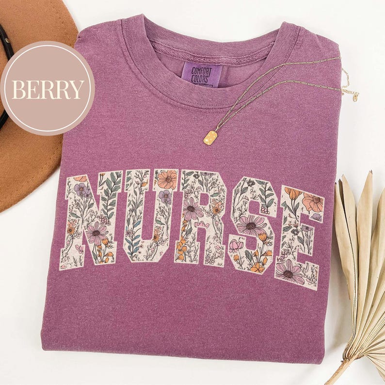 May include: A berry-colored t-shirt with the word "NURSE" in a floral design. The shirt has a crew neck and a small tag that says "COMFORT COLORS". A gold necklace with a rectangular pendant is draped across the shirt.