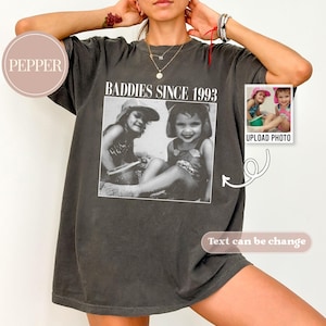 K&ouml;nnte beinhalten: Ein dunkelgraues T-Shirt mit dem Aufdruck "Baddies Since 1993" auf der Vorderseite. Das Shirt zeigt ein Schwarzwei&szlig;foto von zwei M&auml;dchen in Badeanz&uuml;gen. Der Text "Foto hochladen" ist unter dem Foto gedruckt.
