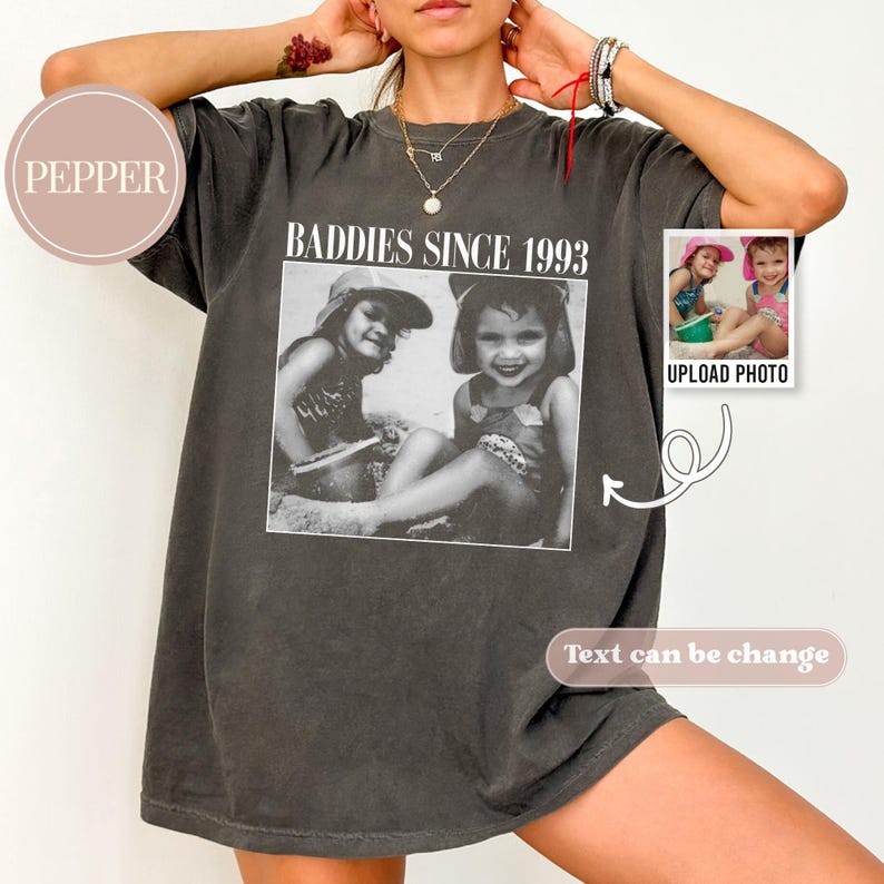 K&ouml;nnte beinhalten: Ein dunkelgraues T-Shirt mit dem Aufdruck "Baddies Since 1993" auf der Vorderseite. Das Shirt zeigt ein Schwarzwei&szlig;foto von zwei M&auml;dchen in Badeanz&uuml;gen. Der Text "Foto hochladen" ist unter dem Foto gedruckt.