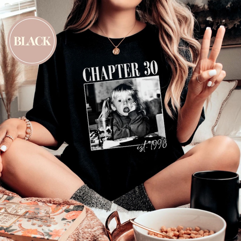 May include: A white ceramic mug with a black handle and the text "But First, Coffee" in a bold, black font. The mug is set against a blurred background, suggesting a cozy setting.
