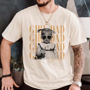 May include: Cream-colored t-shirt featuring a black and white photo of a child wearing heart-shaped sunglasses and making a peace sign. The words "GIRL DAD" are printed in gold, with "Est. 2020" below the photo.