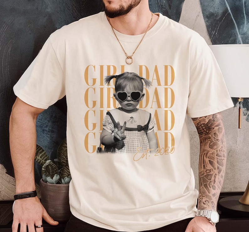 May include: Cream-colored t-shirt featuring a black and white photo of a child wearing heart-shaped sunglasses and making a peace sign. The words "GIRL DAD" are printed in gold, with "Est. 2020" below the photo.
