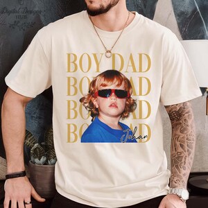 Pode incluir: Camiseta creme com foto de crian&ccedil;a usando &oacute;culos de sol vermelhos e camisa azul. As palavras "BOY DAD" s&atilde;o impressas em dourado verticalmente. O nome da crian&ccedil;a, "Johan", est&aacute; escrito em it&aacute;lico.