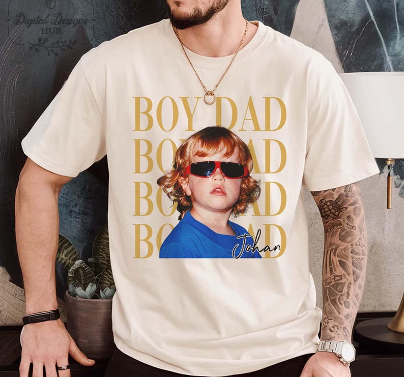Pode incluir: Camiseta creme com foto de crian&ccedil;a usando &oacute;culos de sol vermelhos e camisa azul. As palavras "BOY DAD" s&atilde;o impressas em dourado verticalmente. O nome da crian&ccedil;a, "Johan", est&aacute; escrito em it&aacute;lico.
