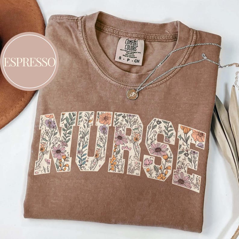 May include: Espresso-colored t-shirt with the word "NURSE" in large, floral-filled letters. The shirt has a crew neck and short sleeves. A silver necklace with a pendant is draped across the shirt. The shirt is folded on a white surface.