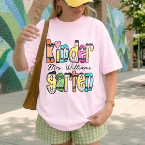 May include: A light pink short-sleeve t-shirt featuring a colorful graphic print with the text Kinder, Mrs. Williams, and garten. The letters are styled to resemble crayons in various colors including orange, blue, pink, yellow, and green.
