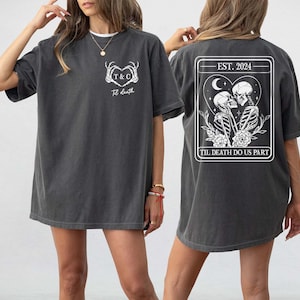 May include: Dark gray t-shirt with a white graphic on the front and back. The front features a heart-shaped design with the text "T & C" and "Til death". The back has a framed design with skeletons kissing, the text "Est. 2024" and "Til death do us part".