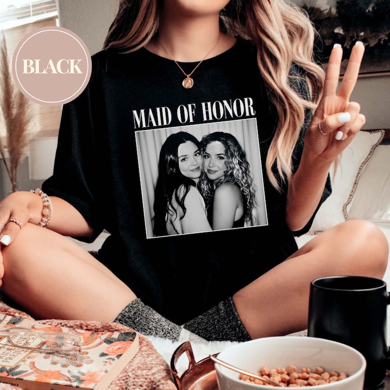 Op de afbeelding: Zwart T-shirt met de tekst "MAID OF HONOR" boven een zwart-witfoto van twee vrouwen. Het shirt wordt gedragen door een persoon die met gekruiste benen zit, met een gouden ketting en armbanden.
