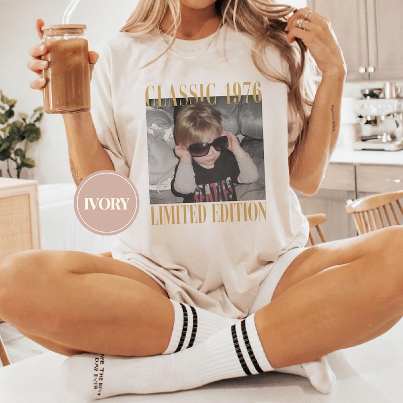 May include: Ivory-colored t-shirt with a vintage photo print. The photo features a child wearing sunglasses, with the text "CLASSIC 1976 LIMITED EDITION" in gold lettering. The shirt is a casual style.