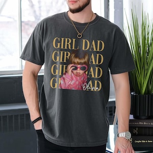 Pode incluir: Camiseta cinza escuro com as palavras "GIRL DAD" em letras douradas. A camiseta apresenta uma foto de uma crian&ccedil;a usando &oacute;culos de sol. O nome da crian&ccedil;a, Olivia, est&aacute; escrito abaixo.
