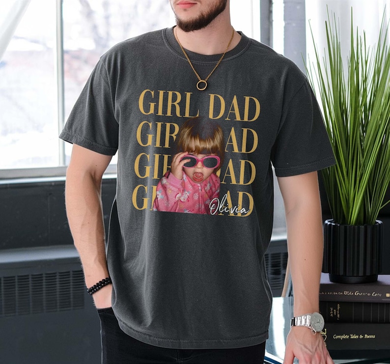 Pode incluir: Camiseta cinza escuro com as palavras "GIRL DAD" em letras douradas. A camiseta apresenta uma foto de uma crian&ccedil;a usando &oacute;culos de sol. O nome da crian&ccedil;a, Olivia, est&aacute; escrito abaixo.