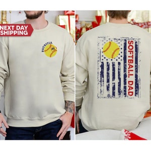 May include: A beige sweatshirt with a distressed American flag design on the back. The flag has a softball in the center and the text "Softball Dad" written vertically. The front of the sweatshirt has a softball with the text "Patricia 24" on it.