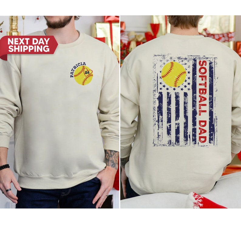 May include: A beige sweatshirt with a distressed American flag design on the back. The flag has a softball in the center and the text "Softball Dad" written vertically. The front of the sweatshirt has a softball with the text "Patricia 24" on it.
