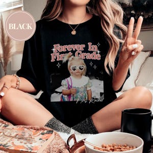K&ouml;nnte beinhalten: Schwarzes T-Shirt mit dem Text "Forever In First Grade" in Rosa und Wei&szlig;, mit einem Foto eines kleinen Kindes mit herzf&ouml;rmiger Sonnenbrille. Der Text "Mrs. Rachel" steht unter dem Foto. Ein schwarzer Kreis mit dem Wort "BLACK" befindet sich oben links.