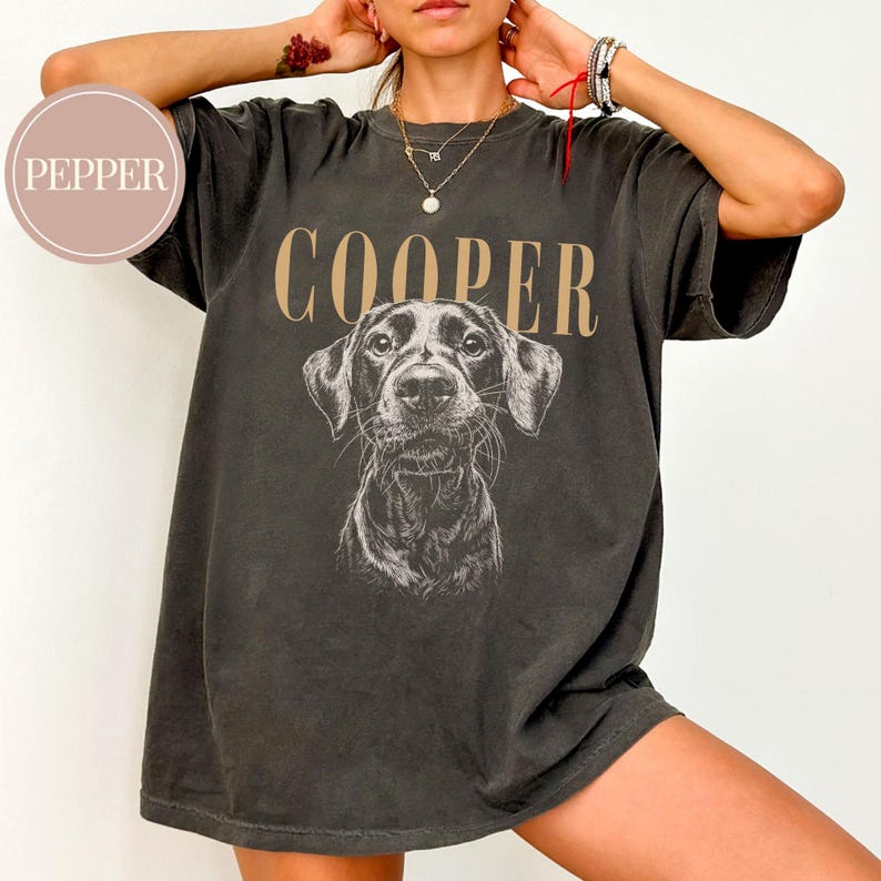 May include: Dark gray t-shirt featuring a detailed illustration of a dog's face, with the name "COOPER" printed above in a tan color. The shirt has a vintage, worn look.