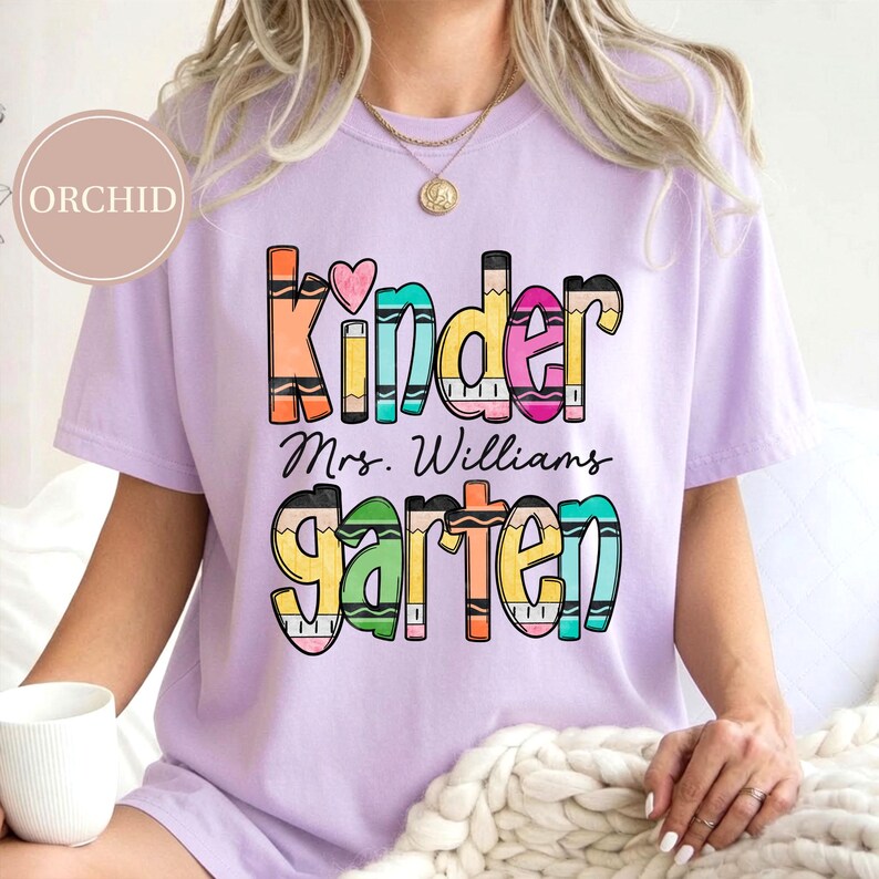 May include: A light purple short-sleeve t-shirt featuring a graphic design with the text Kinder and Garten in a colorful crayon-inspired font. A small pink heart is above the text, and the name Mrs. Williams is printed in black script.