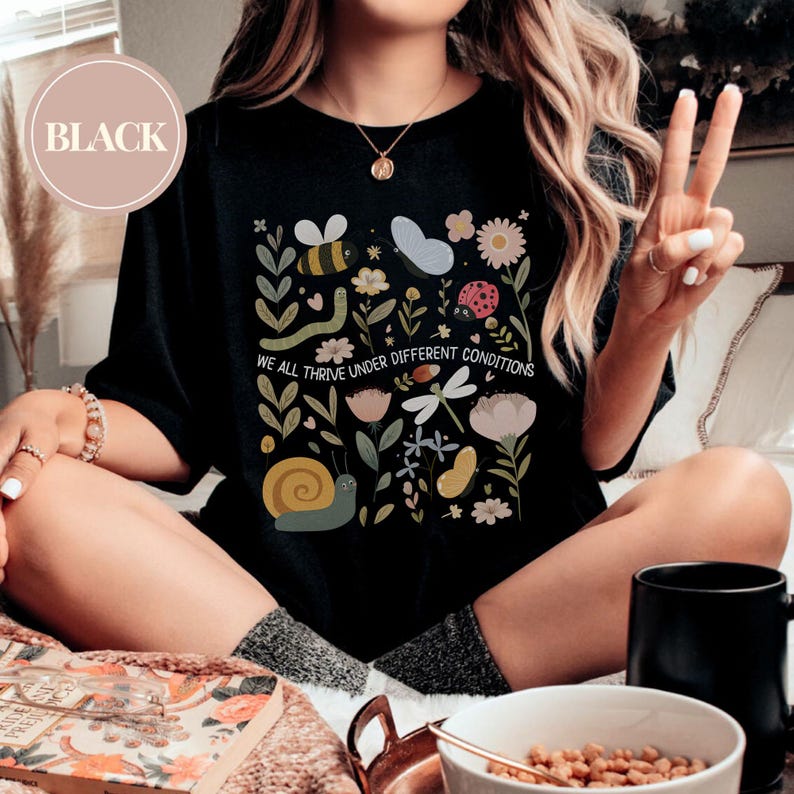 May include: Black crew neck t-shirt with a vibrant graphic of insects and flowers. The text "WE ALL THRIVE UNDER DIFFERENT CONDITIONS" is printed below the graphic. The design includes a bee, butterfly, and ladybug.