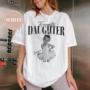 May include: White t-shirt with the words "Favorite Daughter" in black script and a black and white photo of a young girl in a tutu. The name "Emma" is written below the photo. A circular label in the upper left corner reads "WHITE".