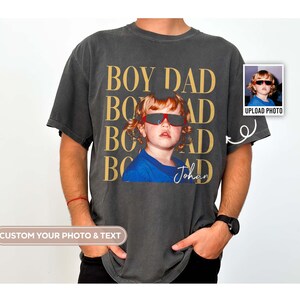 Pode incluir: T-shirt cinzento escuro com as palavras douradas "BOY DAD". A camisa apresenta uma foto de uma crian&ccedil;a usando &oacute;culos de sol. O nome "Johan" est&aacute; escrito abaixo da foto. Uma pequena foto est&aacute; no canto superior direito com o texto "UPLOAD PHOTO".