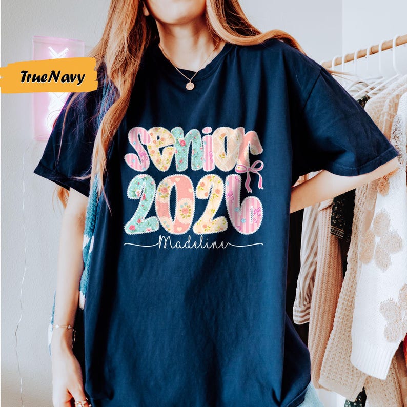 May include: Navy blue t-shirt with the text "Senior 2026 Madeline" in a colorful, floral design. The shirt has a relaxed fit and is suitable for graduation celebrations.
