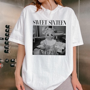 May include: White t-shirt featuring a black and white photograph of a young child wearing heart-shaped sunglasses. The text "SWEET SIXTEEN" is printed above the image in a black, bold font. The shirt has short sleeves.