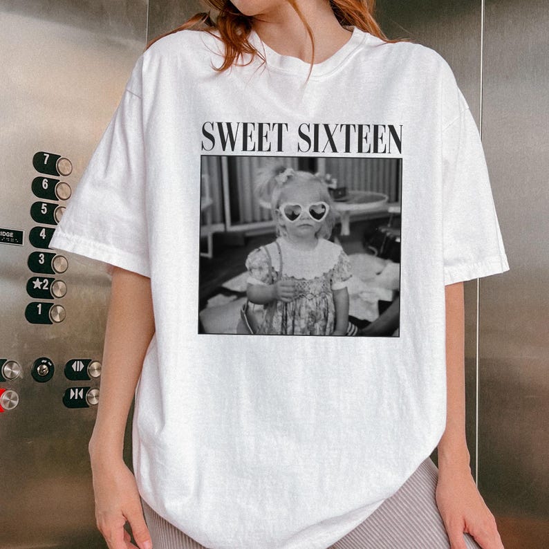 May include: White t-shirt featuring a black and white photograph of a young child wearing heart-shaped sunglasses. The text "SWEET SIXTEEN" is printed above the image in a black, bold font. The shirt has short sleeves.