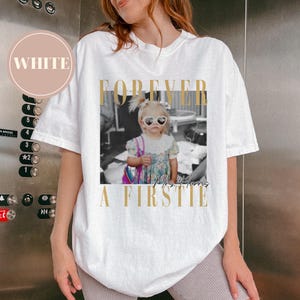 May include: White t-shirt with a graphic featuring a black and white photo of a young girl wearing heart-shaped sunglasses. The text "FOREVER A FIRSTIE Mrs. Harris" is printed in gold.