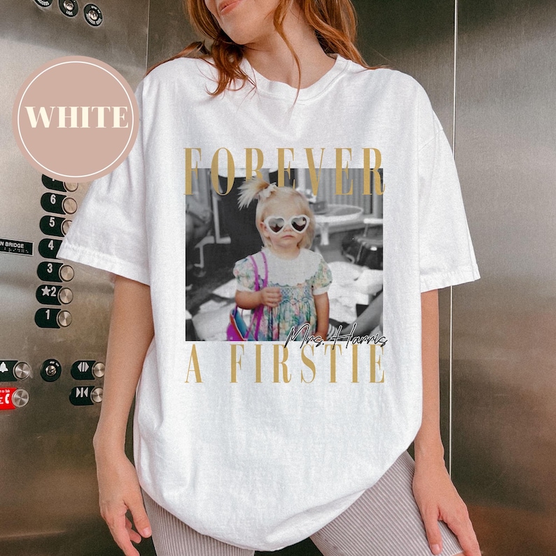 May include: White t-shirt with a graphic featuring a black and white photo of a young girl wearing heart-shaped sunglasses. The text "FOREVER A FIRSTIE Mrs. Harris" is printed in gold.