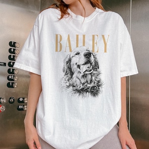 May include: White t-shirt featuring a detailed black and white illustration of a Golden Retriever dog with its tongue out. The word "BAILEY" is printed in gold above the dog image. The shirt has short sleeves.