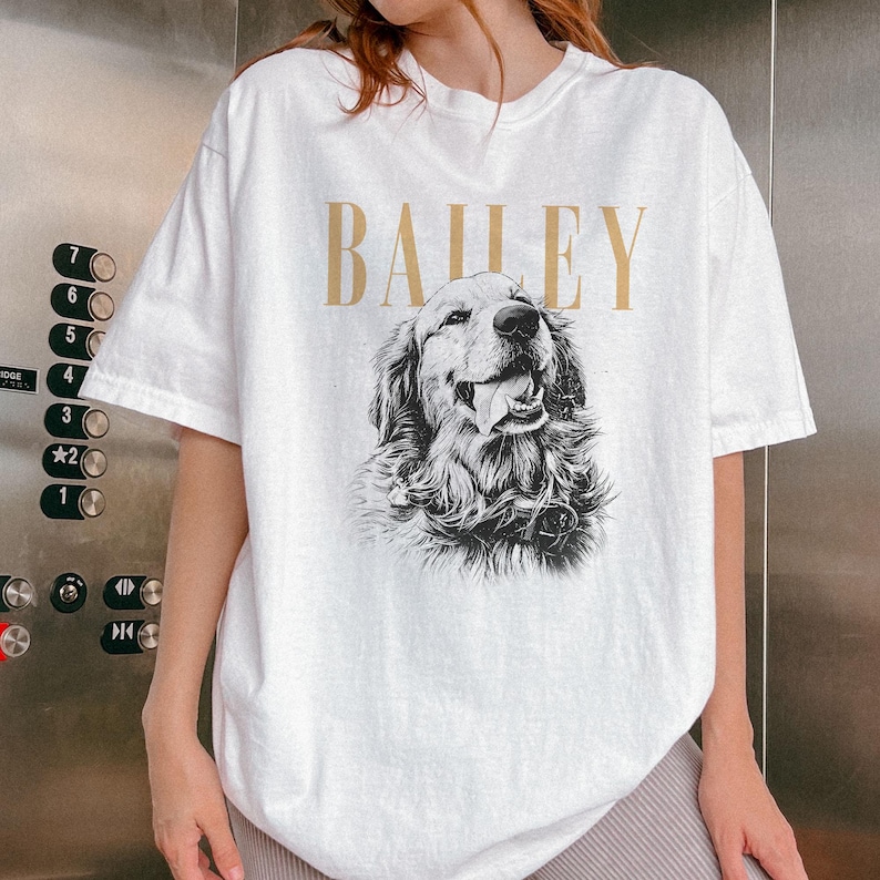 May include: White t-shirt featuring a detailed black and white illustration of a Golden Retriever dog with its tongue out. The word "BAILEY" is printed in gold above the dog image. The shirt has short sleeves.