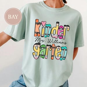May include: A light green short-sleeve t-shirt featuring a graphic print with the text "Kinder" and "garten" in colorful, crayon-inspired lettering, with "Mrs. Williams" written in black script between the two words and a small pink heart.