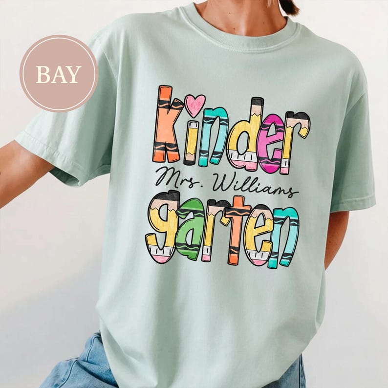 May include: A light green short-sleeve t-shirt featuring a graphic print with the text "Kinder" and "garten" in colorful, crayon-inspired lettering, with "Mrs. Williams" written in black script between the two words and a small pink heart.