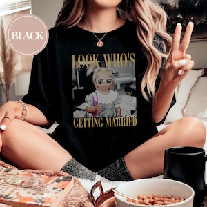 May include: Black t-shirt with a gold-colored graphic that says "LOOK WHO'S GETTING MARRIED" with a black and white photo of a young girl wearing heart-shaped sunglasses. The word "Lauren" is written below the photo.