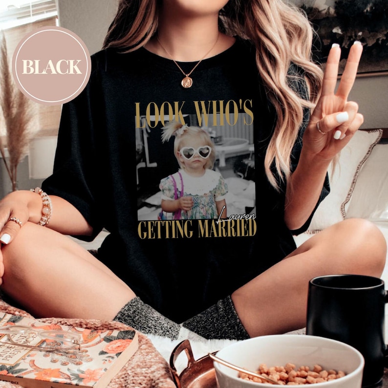 May include: Black t-shirt with a gold-colored graphic that says "LOOK WHO'S GETTING MARRIED" with a black and white photo of a young girl wearing heart-shaped sunglasses. The word "Lauren" is written below the photo.