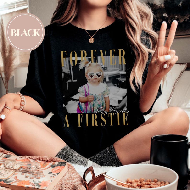 May include: Black t-shirt with a gold graphic that says "FOREVER A FIRSTIE" and a black and white photo of a young girl wearing heart-shaped sunglasses. The text "Mrs. Harris" is also on the graphic.