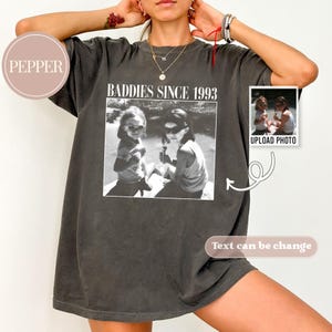 K&ouml;nnte beinhalten: Ein dunkelgraues T-Shirt mit dem Aufdruck "Baddies Since 1993" auf der Vorderseite. Das Shirt zeigt ein Schwarzwei&szlig;foto von zwei M&auml;dchen mit Sonnenbrillen. Der Text "Foto hochladen" ist unter dem Foto gedruckt.