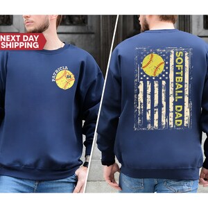 May include: Navy blue sweatshirt with a distressed American flag design and the text "Softball Dad" on the back. The front of the sweatshirt features a softball with the text "Patricia 24" on it.