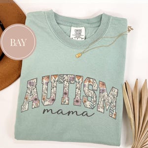 May include: A light green t-shirt with a floral print of the word "AUTISM" and the text "mama" underneath.