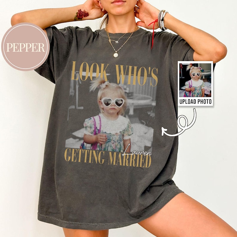 May include: Dark gray t-shirt with a photo of a young girl wearing heart-shaped sunglasses. The text "LOOK WHO'S GETTING MARRIED" is printed in gold lettering. A small photo with the text "UPLOAD PHOTO" is on the right side.