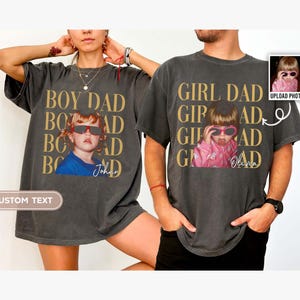 Pode incluir: Duas camisetas cinza escuro com foto e texto personalizados. Uma camisa diz "BOY DAD" com uma foto de uma crian&ccedil;a usando &oacute;culos de sol e o nome "Johan". A outra diz "GIRL DAD" com uma foto de uma crian&ccedil;a e o nome "Olivia".