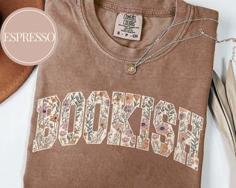 May include: Espresso-colored t-shirt with the word "BOOKISH" in a floral design. The shirt has a crew neck and short sleeves. A silver necklace with a pendant is draped across the shirt. The shirt is folded on a white surface.