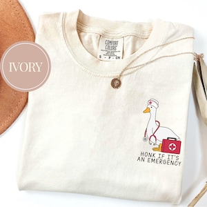 May include: Ivory t-shirt with a cartoon illustration of a goose wearing a nurse's hat and holding a stethoscope. The text "Honk if it's an emergency" is printed below the goose.