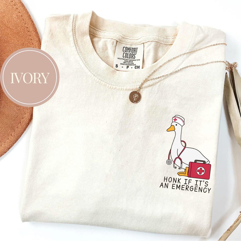 May include: Ivory t-shirt with a cartoon illustration of a goose wearing a nurse's hat and holding a stethoscope. The text "Honk if it's an emergency" is printed below the goose.