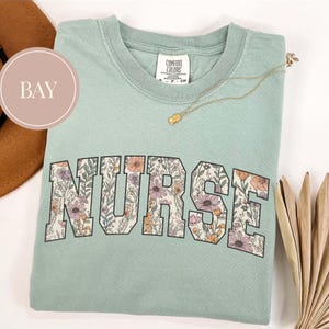 May include: A light green Comfort Colors t-shirt with the word "NURSE" in large, floral-filled letters. A gold necklace with a small pendant rests on the shirt. The shirt is folded on a white surface.