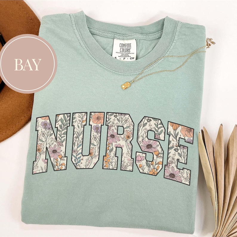 May include: A light green Comfort Colors t-shirt with the word "NURSE" in large, floral-filled letters. A gold necklace with a small pendant rests on the shirt. The shirt is folded on a white surface.