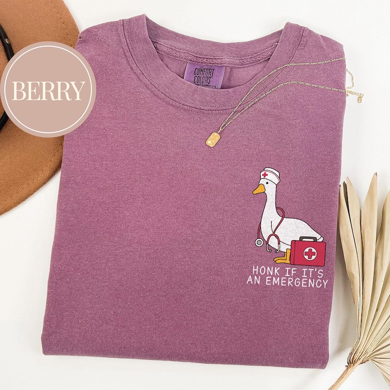 May include: A purple t-shirt with a white goose wearing a nurse's hat and holding a stethoscope and a first aid kit. The text "Honk if it's an emergency" is printed below the goose.