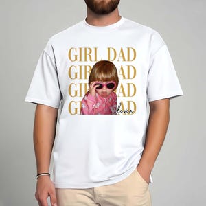 Pode incluir: Camiseta branca com foto de uma crian&ccedil;a usando &oacute;culos de sol rosa e jaqueta rosa. As palavras "GIRL DAD" s&atilde;o impressas em dourado acima da foto. O nome da crian&ccedil;a, "Olivia", est&aacute; escrito em it&aacute;lico.