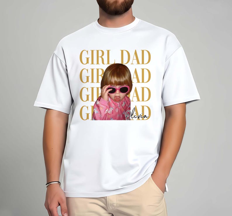 Pode incluir: Camiseta branca com foto de uma crian&ccedil;a usando &oacute;culos de sol rosa e jaqueta rosa. As palavras "GIRL DAD" s&atilde;o impressas em dourado acima da foto. O nome da crian&ccedil;a, "Olivia", est&aacute; escrito em it&aacute;lico.