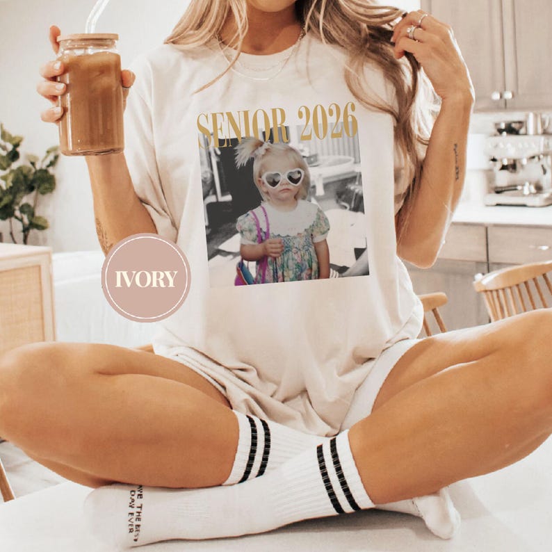 May include: Ivory-colored t-shirt with the text "SENIOR 2026" in gold lettering. The shirt features a black and white photo of a child wearing heart-shaped sunglasses. The person is wearing white socks with black stripes.