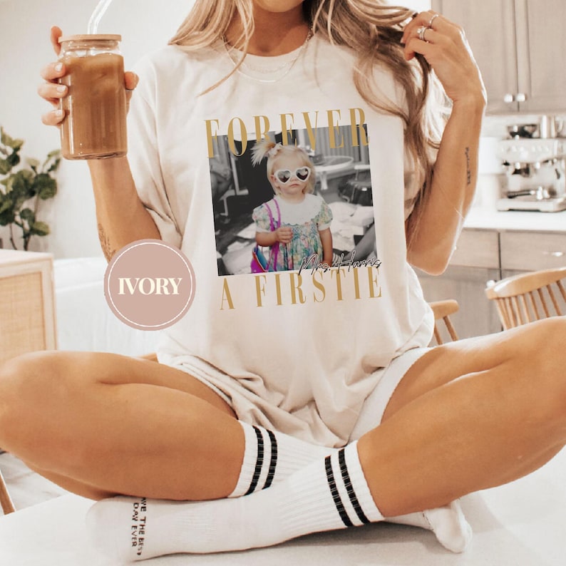 May include: Ivory-colored t-shirt featuring a black and white photo of a child wearing heart-shaped sunglasses. The shirt has the words "FOREVER A FIRSTIE" printed in gold. The person is wearing white socks with black stripes.